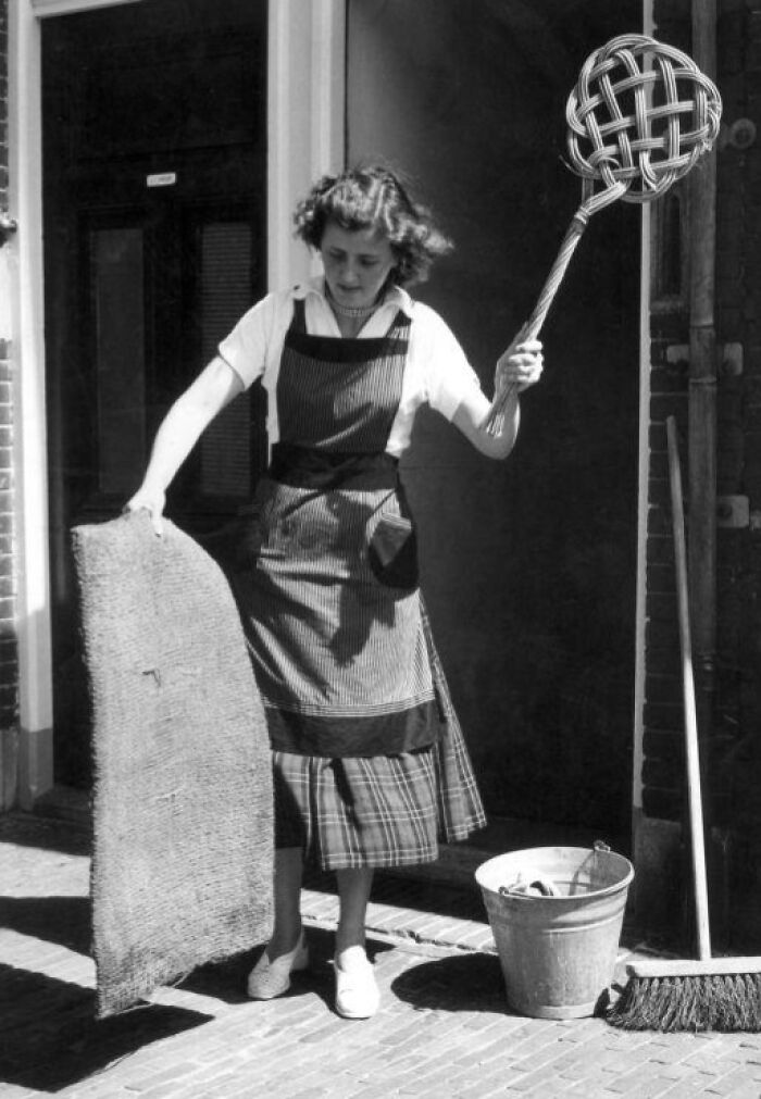 1940s housewife in apron cleaning outside with broom, bucket, and mat, showcasing iconic housewife roles of the era.