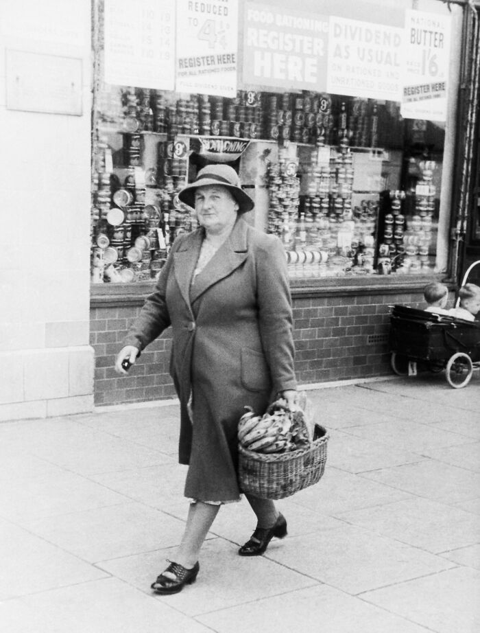 1940s housewife walking with a basket of groceries in front of a shop window during wartime rationing.