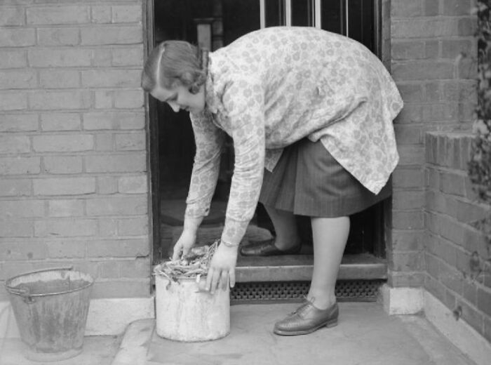 1940s housewife bending to place sticks in a bucket outside a brick house doorway in black and white photo