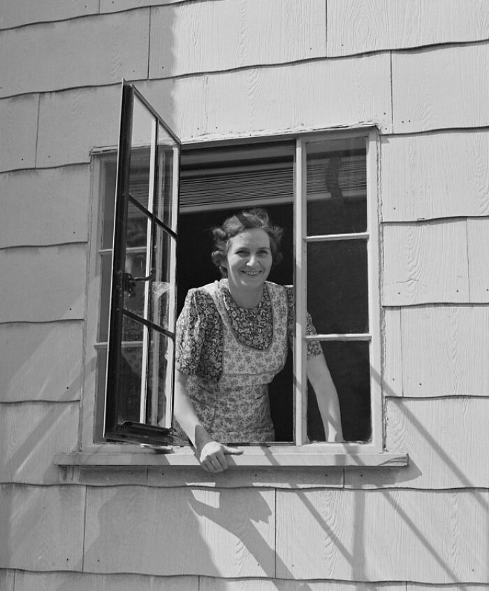 1940s housewife smiling and leaning out of an open window of a wood-paneled home on a sunny day.