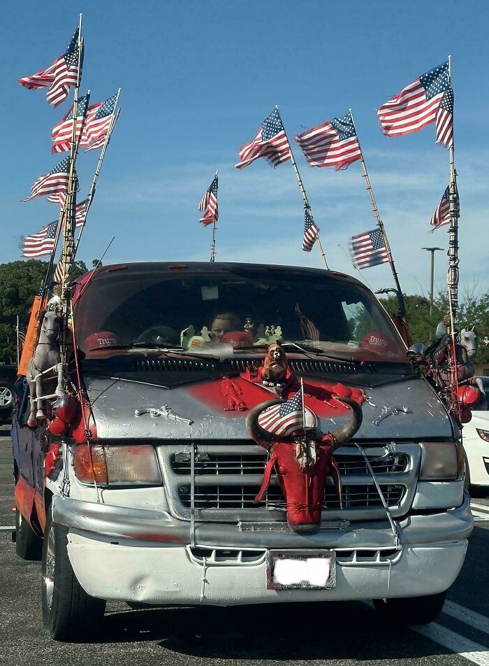 Van with outrageous car mods including multiple American flags and a decorated bull skull on the hood in a parking lot.
