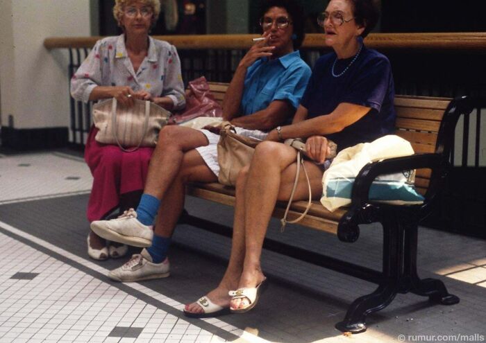 Three older women sitting on a bench in a mall, one smoking, showcasing a ridiculous pic from the past.