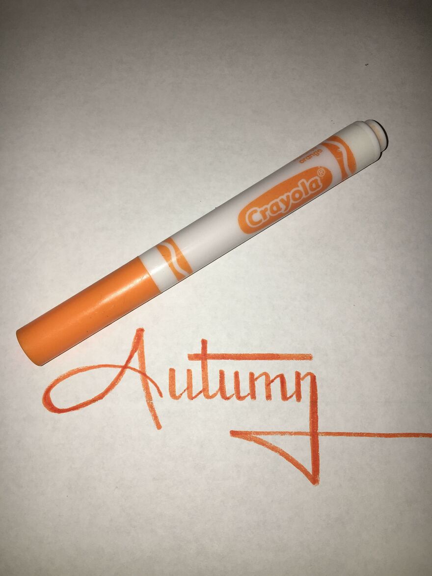 Orange Crayola marker placed above perfect handwriting example of the word Autumn on white paper.