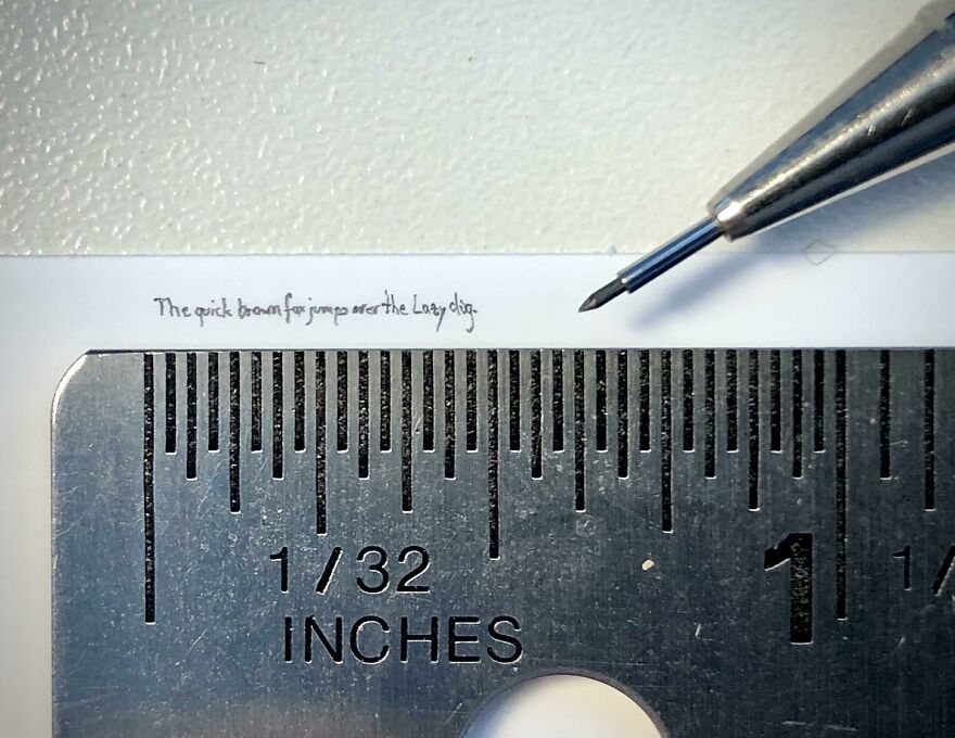Extremely small perfect handwriting example measured against a metal ruler with a pen tip nearby for scale.