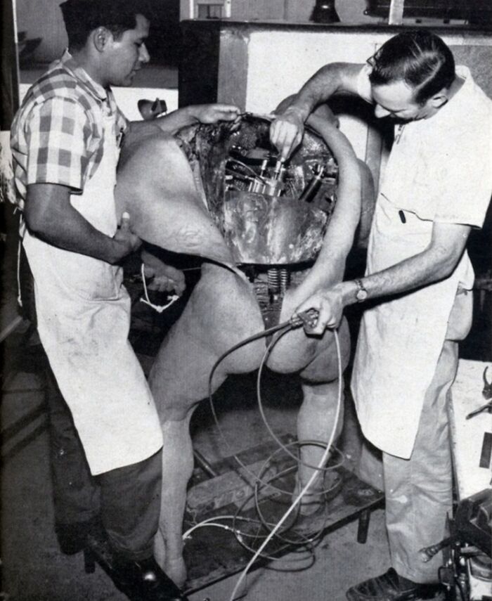 Two men working on an unsettling vintage humanoid robot with exposed mechanical parts and cables in a workshop.