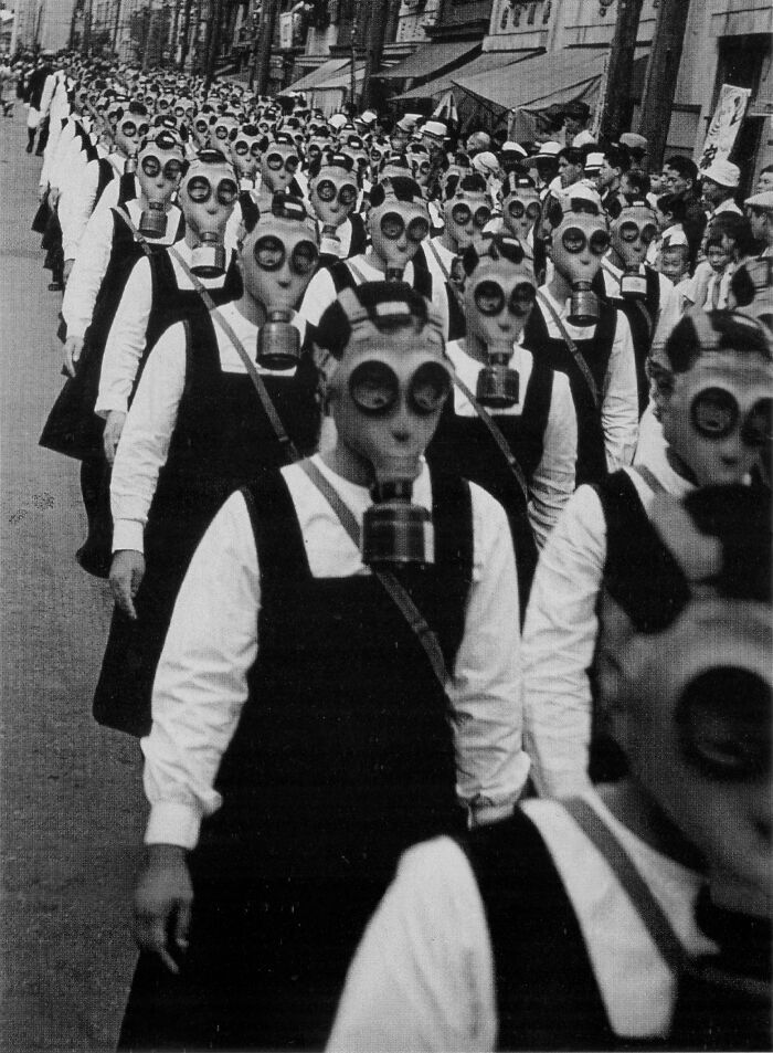 Rows of schoolgirls wearing gas masks march down a street in a creepy and unsettling historic photo.