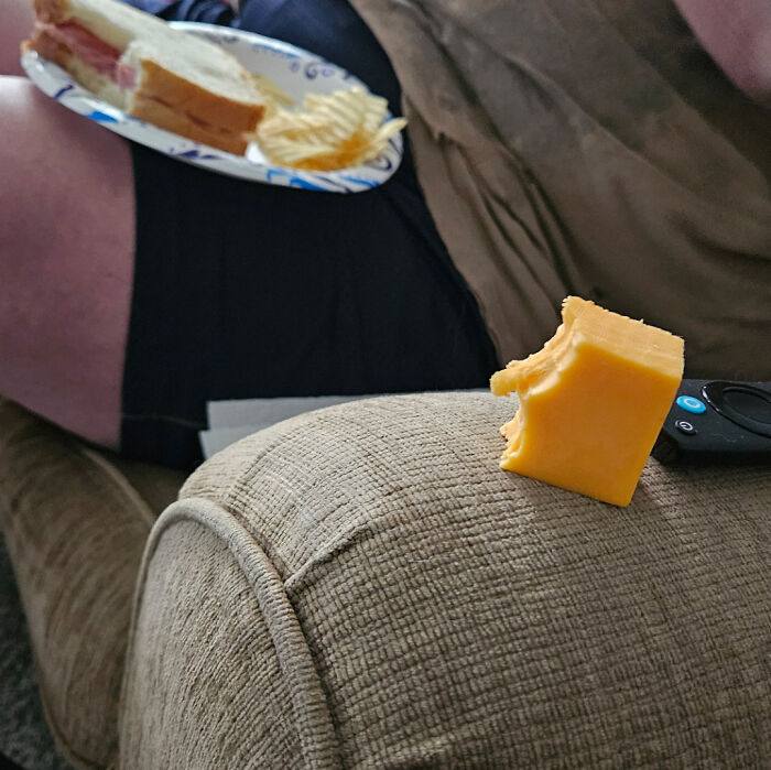 A partially eaten block of cheese left on a couch armrest with a sandwich and chips on a plate nearby showing clueless partner behavior.