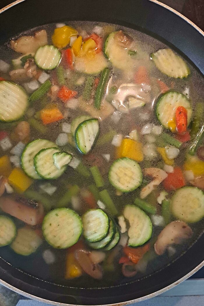 Vegetables including zucchini, bell peppers, and mushrooms cooking in a pan with liquid, showing clueless partner cooking fail.