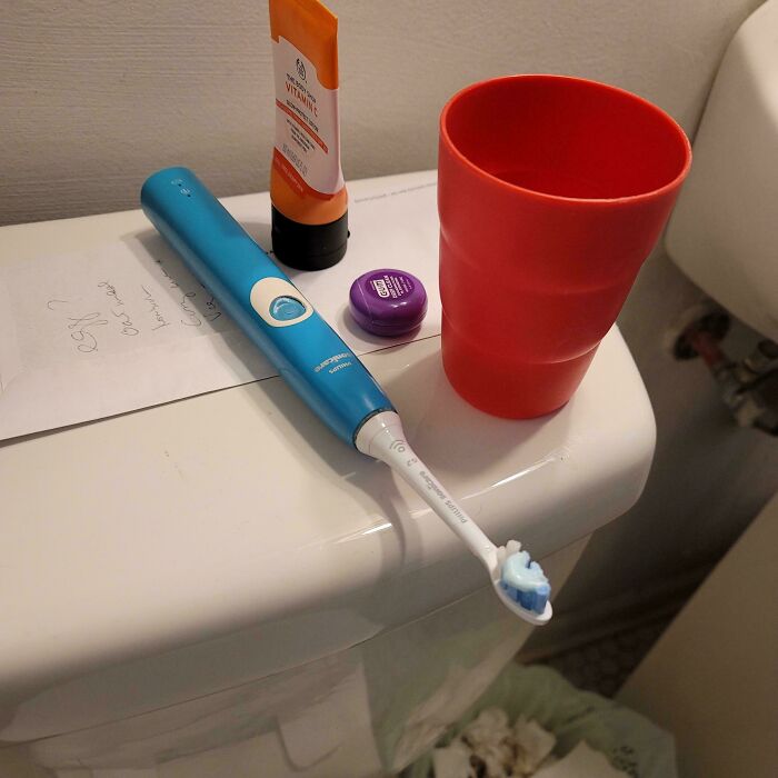 Electric toothbrush left on top of a toilet tank next to toothpaste and a red cup, showing clueless partner behavior.