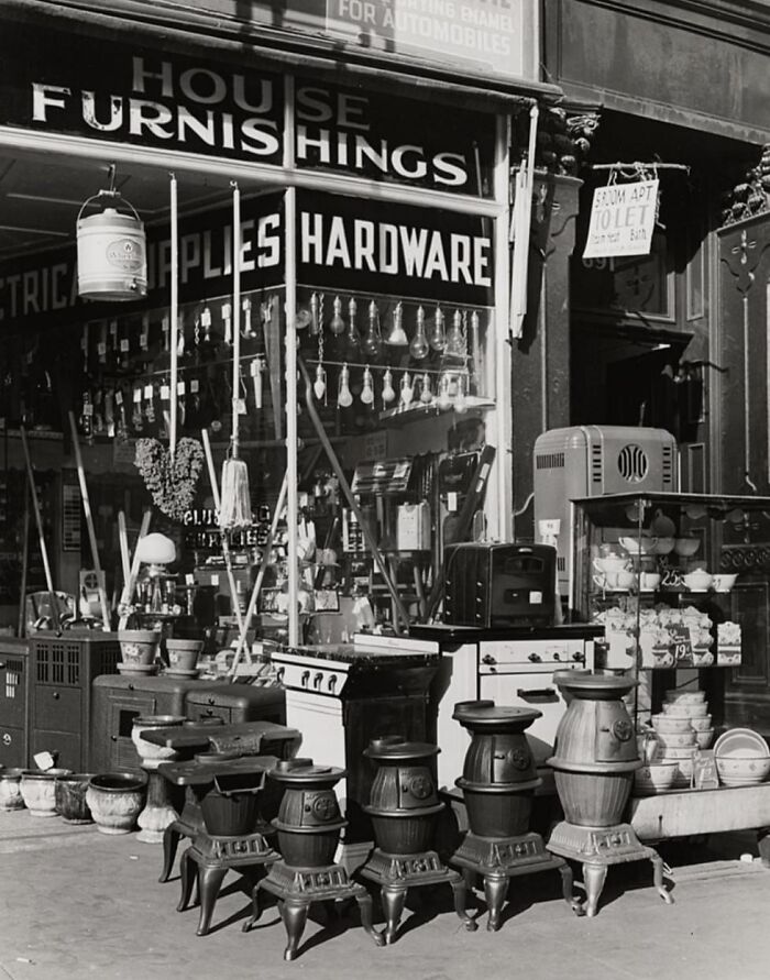 Black and white historical photo of a vintage hardware store displaying stoves, household items, and lighting fixtures outside.