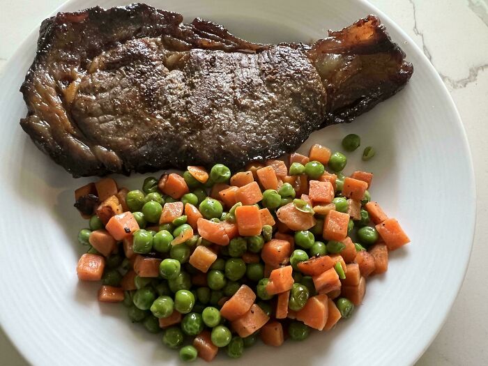 Cooked steak served with mixed peas and carrots on a white plate showing clueless partner meal preparation.