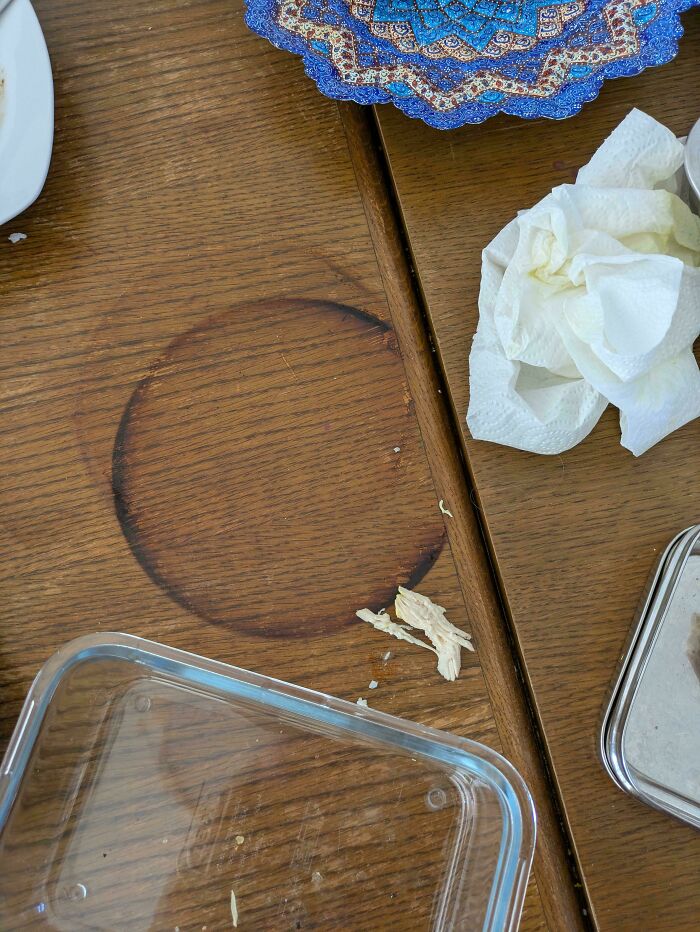 Wooden table with a large coffee ring stain, crumpled napkins, and plastic container showing clueless partner mess.