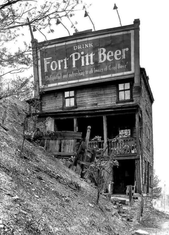 Black and white historical photo of an old wooden building with a Fort Pitt Beer advertisement sign.
