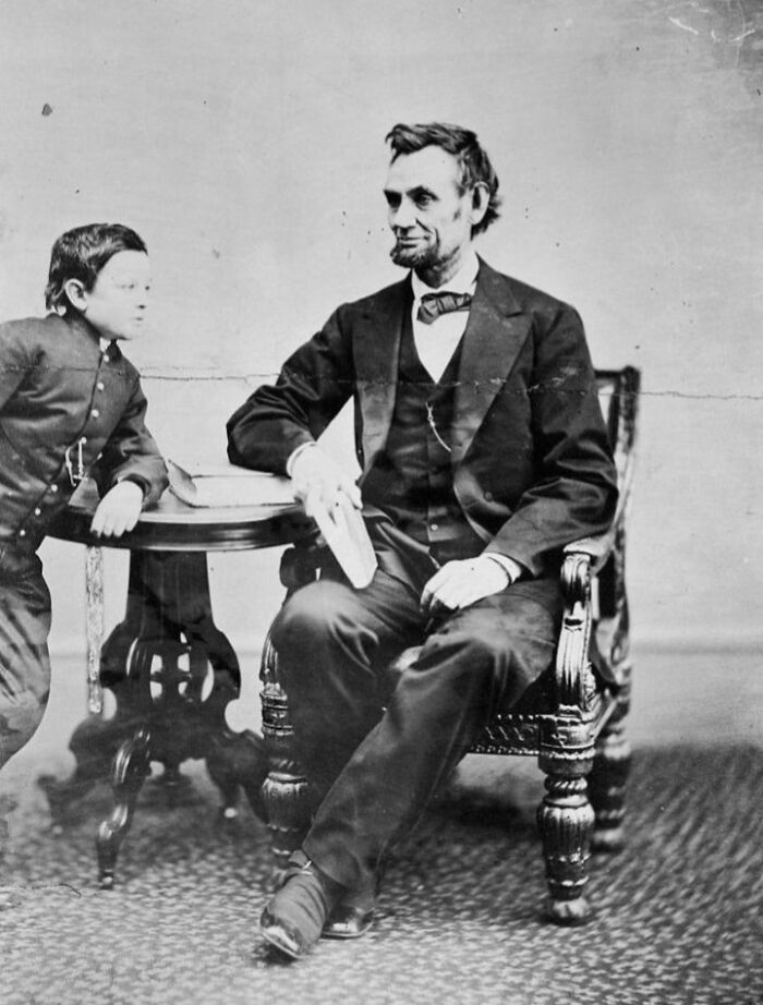 Black and white historical photo showing Abraham Lincoln seated with a young boy leaning on a table beside him.