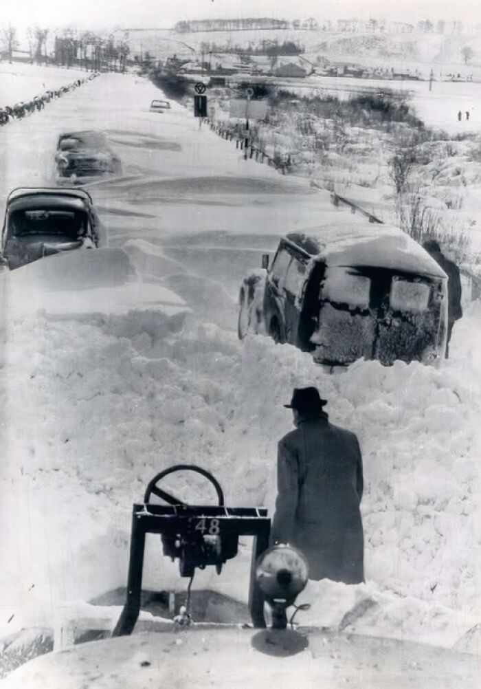 Man in a hat standing by snow-covered vintage cars on a blocked road in a historical winter scene with deep snowdrifts.