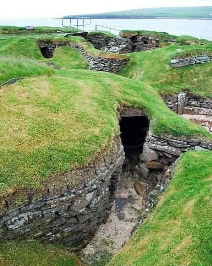 Ancient stone and grass-covered archaeological site with underground chambers near a body of water in archaeology world.