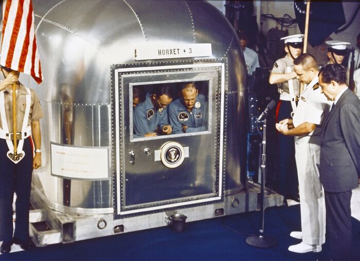 Apollo mission astronauts inside quarantine capsule with military and officials nearby during post-mission procedure.