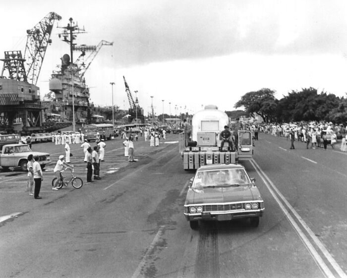 Historic Apollo mission convoy drives through a crowds gathering at a busy shipyard dock beside cranes and ships.