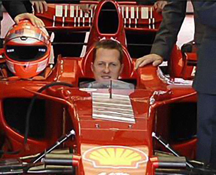 Michael Schumacher sitting in a red Formula 1 car, with his helmet beside him and a person resting hand on the car.