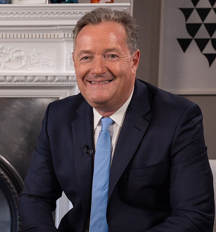 Man in a dark suit and blue tie sitting and smiling indoors, representing famous people who did evil things ignored by the world.