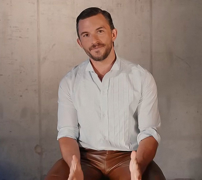 Man in a white shirt and brown pants sitting against a plain wall, posing with hands slightly raised and smiling.