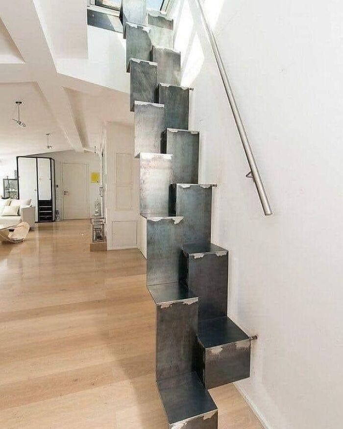 Narrow, steep metal death stairs with alternating steps and a handrail in a modern, bright living space.