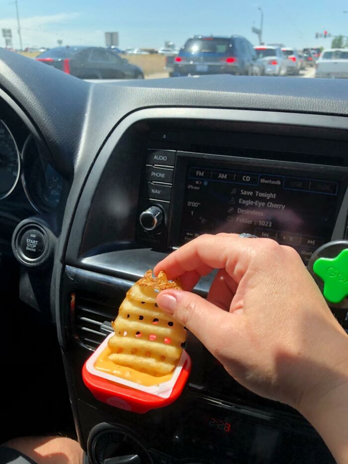 Hand holding a waffle fry dipped in sauce using a clever car find snack holder attached to the dashboard vent.