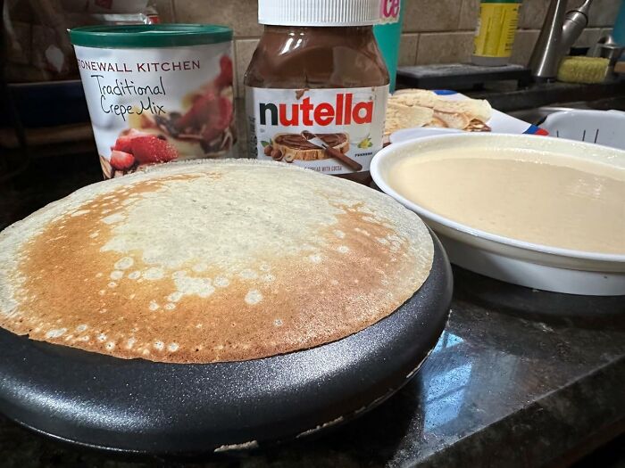 Crepe cooking on a nonstick pan with traditional crepe mix and Nutella jar nearby, showcasing kitchen gadgets.