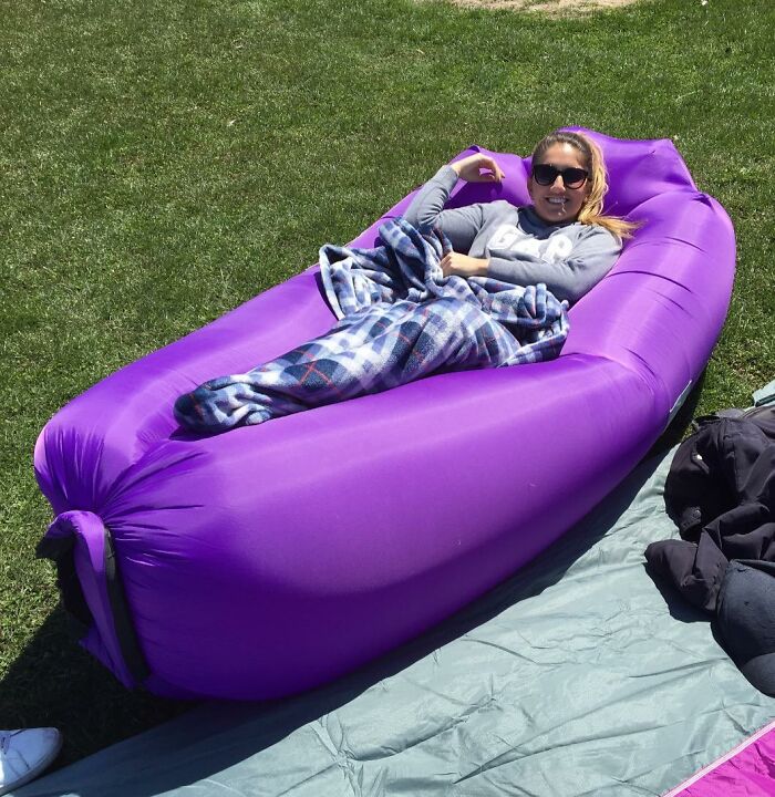 Woman relaxing on a purple inflatable lounger, enjoying a beach day with sun, sand, and snack essentials outdoors.