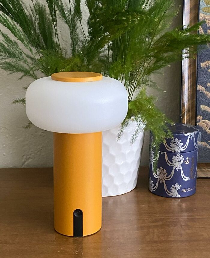 Modern yellow outdoor lamp with white frosted top placed on wooden table near green plant and decorative container.