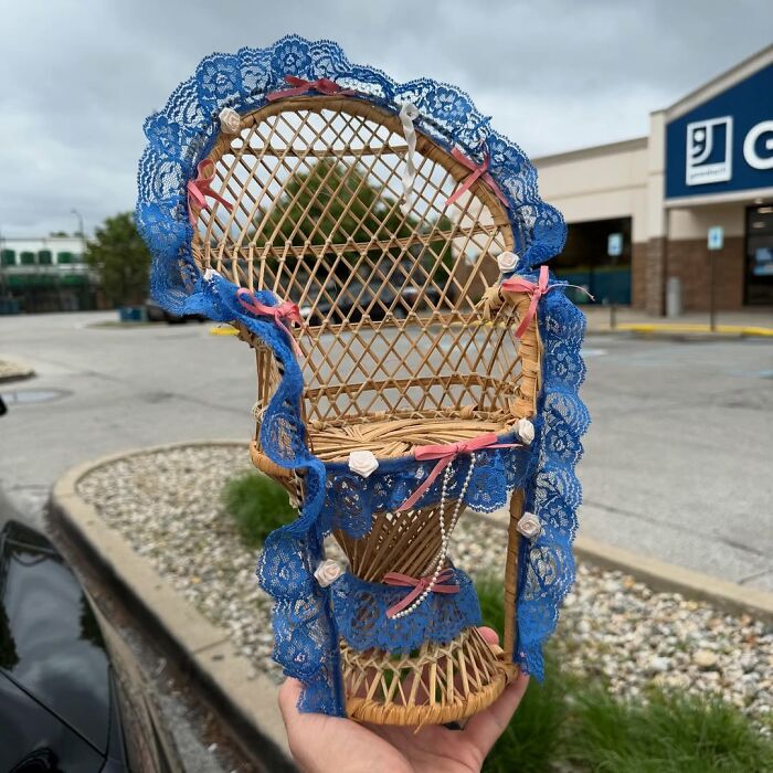 Wicker chair decorated with blue lace and pink ribbons, a cool secondhand find displayed outdoors near a store parking lot.