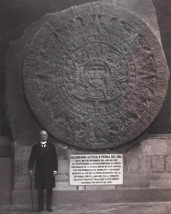 Man standing beside the large Aztec calendar stone, showcasing ancient archaeology world artifact in a museum setting.