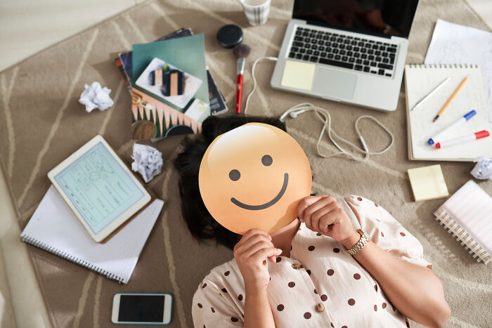 Person lying on floor surrounded by work materials hiding face behind a smiley sign, highlighting small signs of difficult childhood.