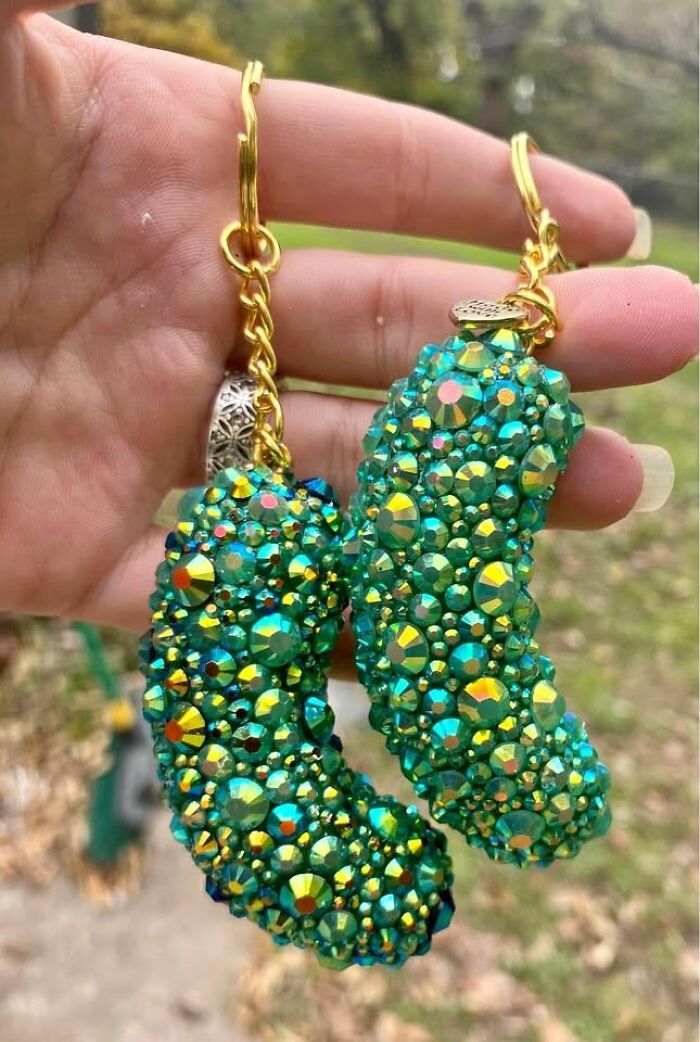 Hand holding DIY bejeweled earrings shaped like pickles, covered in green and gold rhinestones outdoors.
