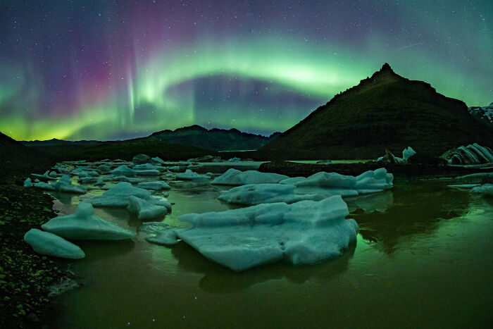 Northern lights over icy landscape with mountains, captured in breathtaking travel photos by the Bored Panda community.