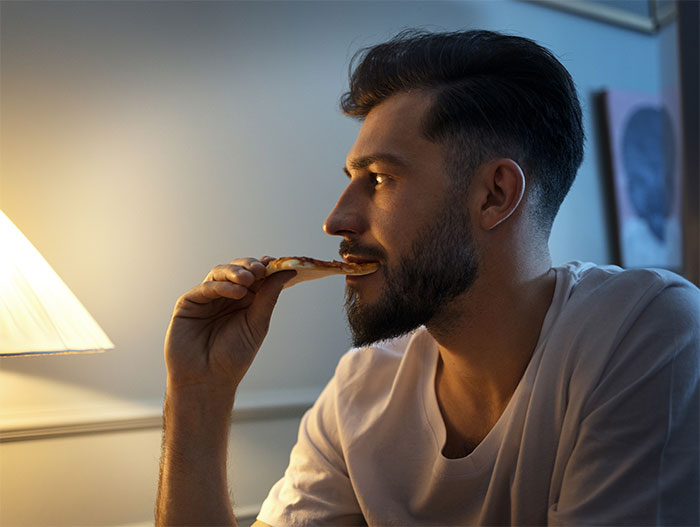 Young man eating pizza alone in dimly lit room, reflecting on years of neglect and family struggles.
