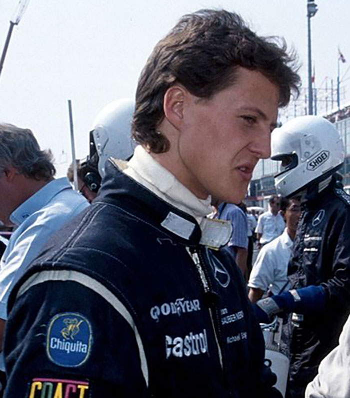 Michael Schumacher in racing suit at F1 event, with team members and helmets in the background.
