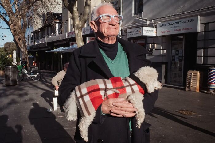 Elderly man wearing glasses holds small dog in a plaid coat on busy city street in candid street photo.