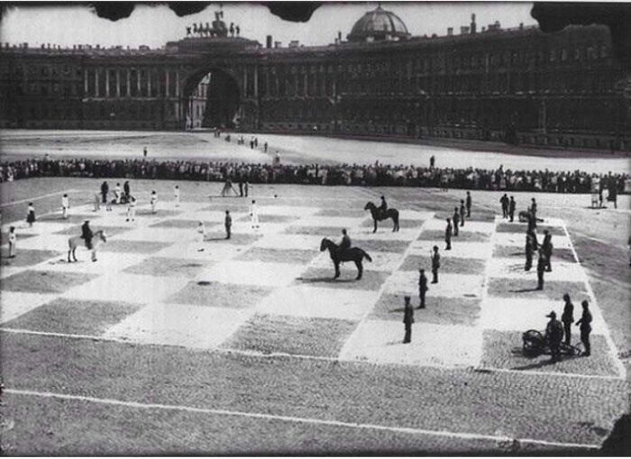 Black and white historical photo of a giant outdoor chess game with people and horses as pieces in a large plaza.