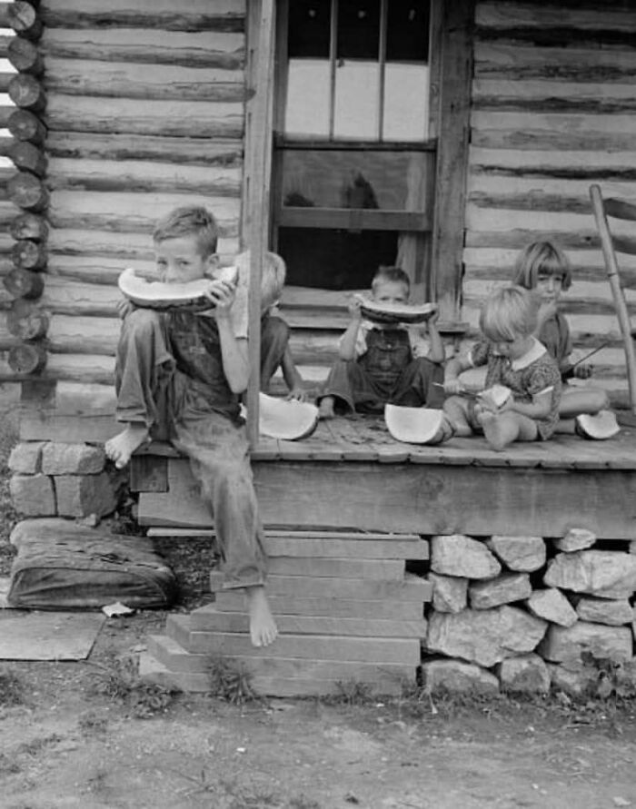 Children sitting on a wooden porch eating watermelon in a historical photo capturing a moment from the past.