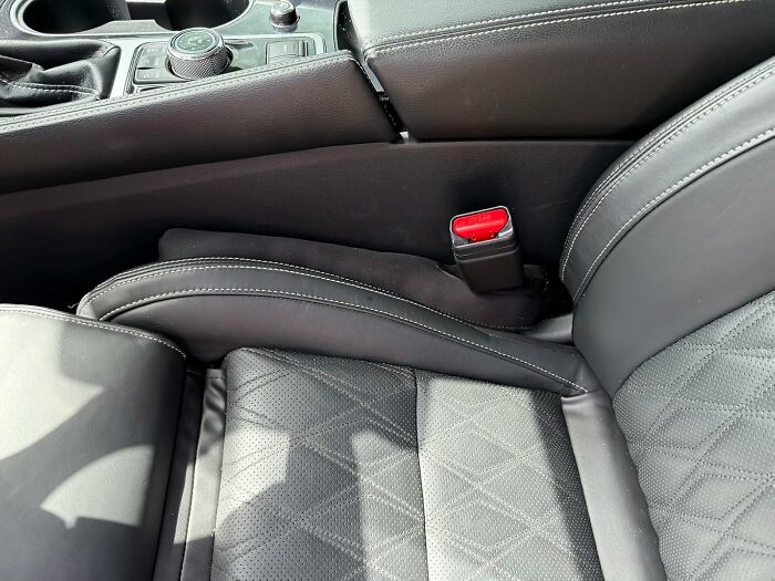 Close-up of black leather car seat and center console highlighting detailed stitching and seatbelt buckle in sunlight.