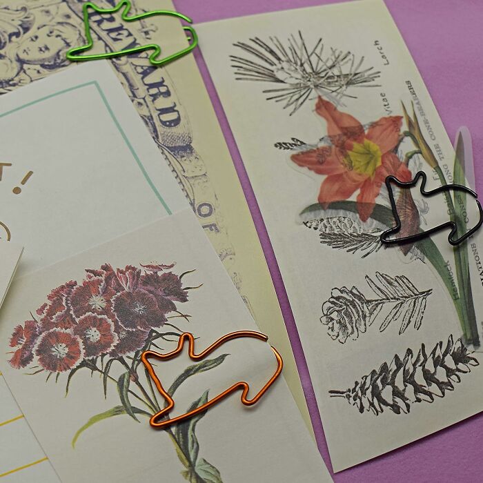 Cute animal-shaped office supplies paper clips holding botanical and vintage-style papers on a purple surface.