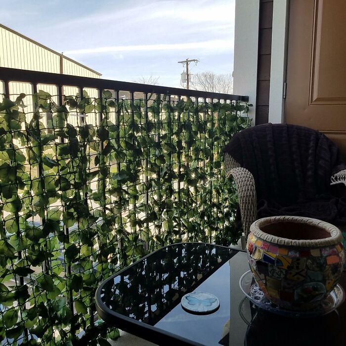 Balcony railing covered with artificial leaves to hide eyesores, featuring a glass table and wicker chair with a dark blanket.