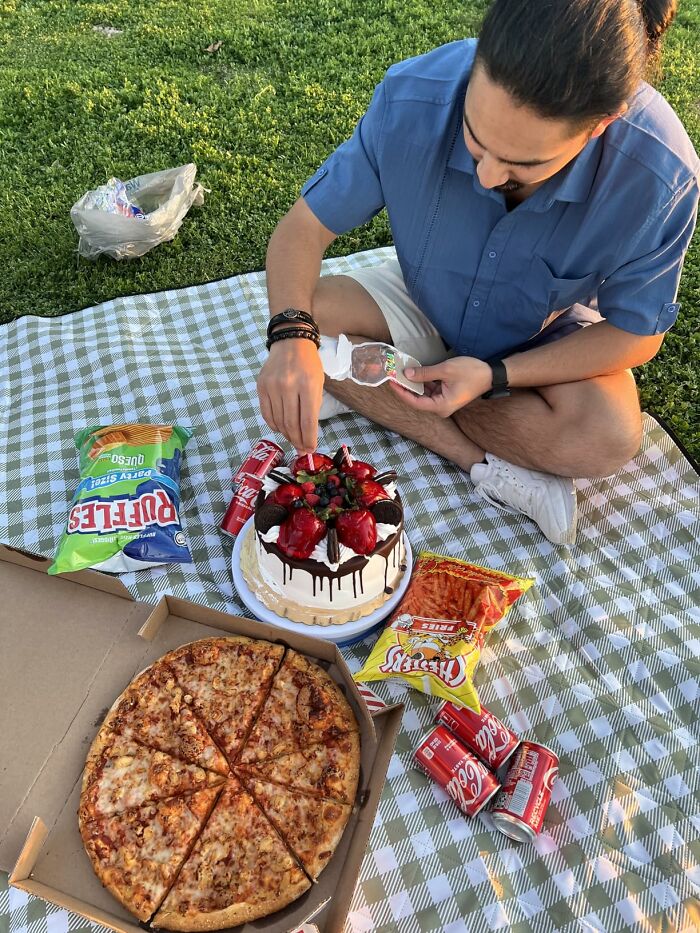 Person sitting on a picnic blanket with pizza, snacks, cake, and sodas for an international picnic day feast outdoors.