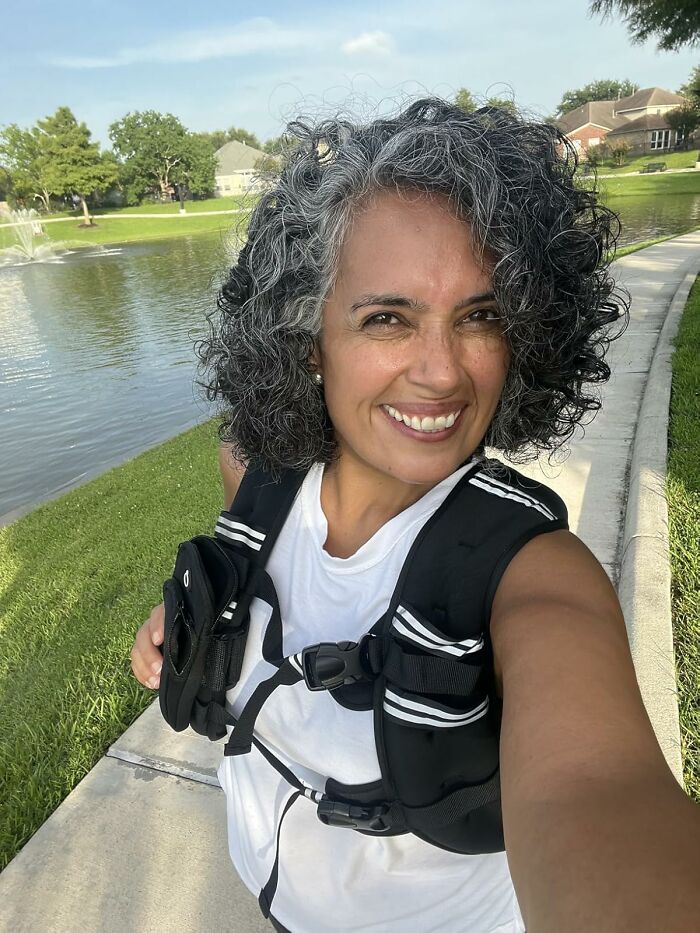 Smiling woman with curly salt-and-pepper hair wearing a black vest outdoors by a lake and sidewalk on a sunny day.