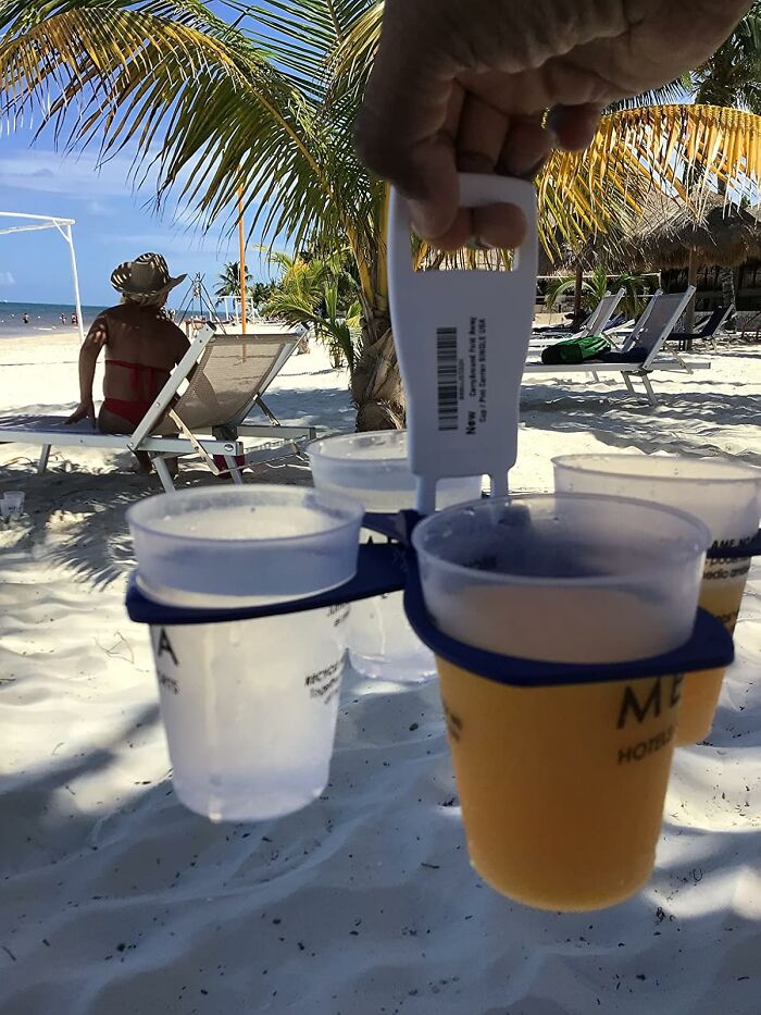 Hand holding a drink carrier with cups on a sandy beach under palm trees, perfect for conquering sun and snack attacks.