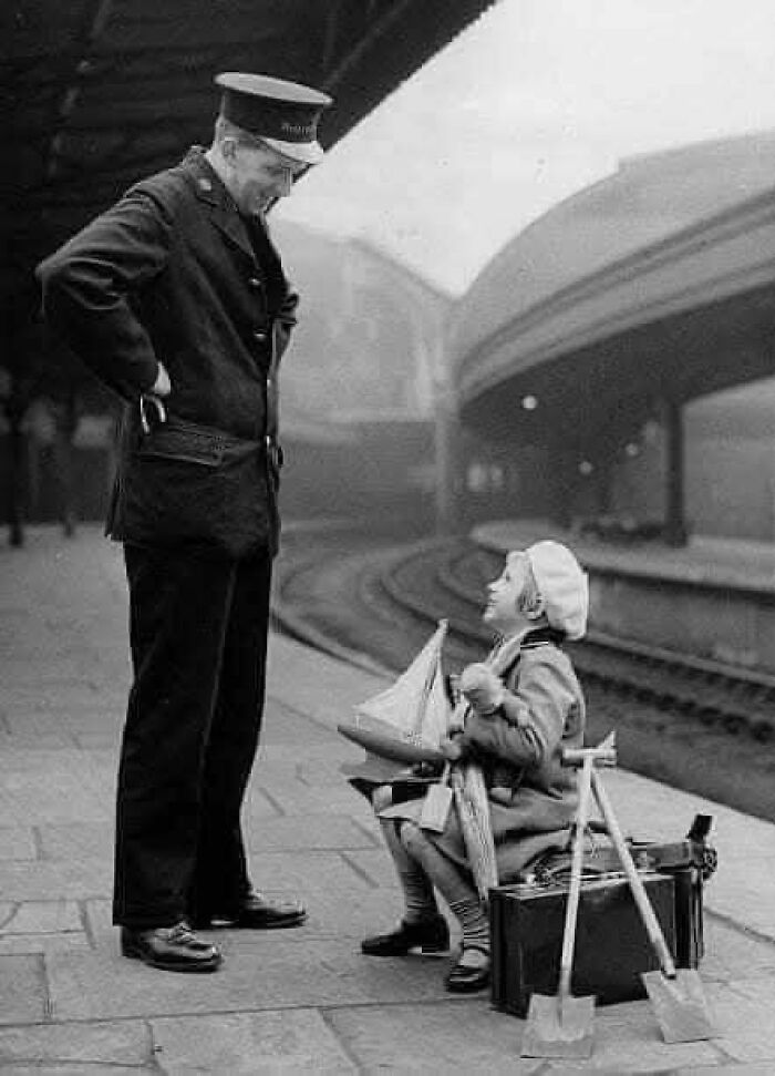 Vintage historical photo of a child holding a toy boat talking to a railway conductor at an empty train station platform.