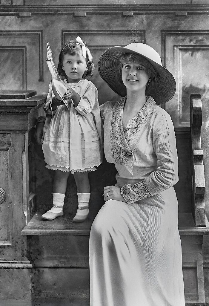 Black and white historical photo of a woman in a large hat with a young child holding a toy boat indoors.