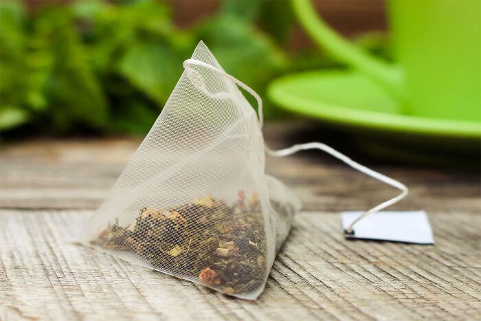Close-up of a pyramid tea bag filled with loose herbs on a wooden surface near a green cup and saucer.