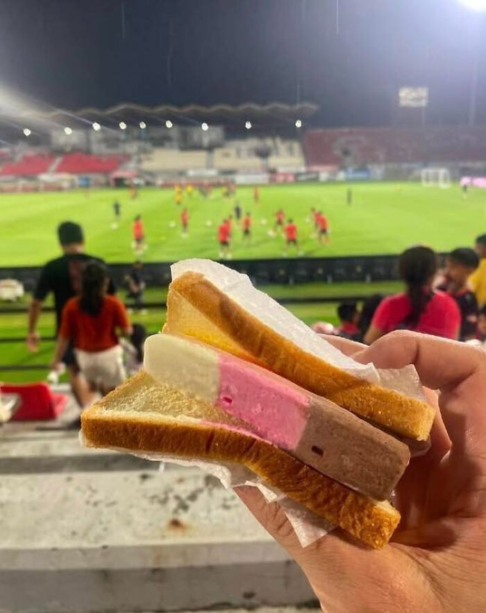 Hand holding an ice cream sandwich made with bread instead of a typical cone, a cursed picture of food at a stadium.